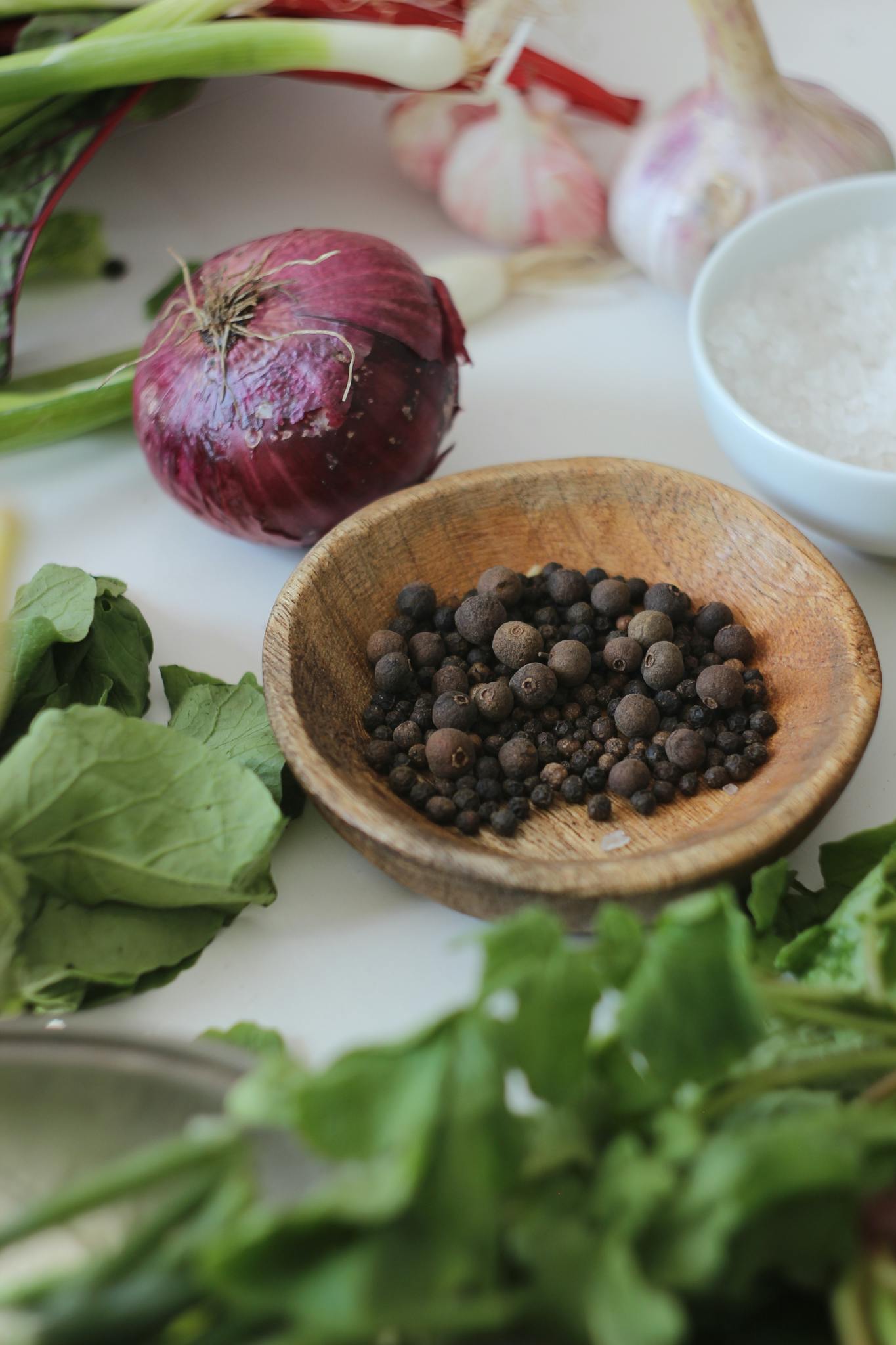 A vertical still life of fresh vegetables and spices including peppercorns, garlic, and herbs.