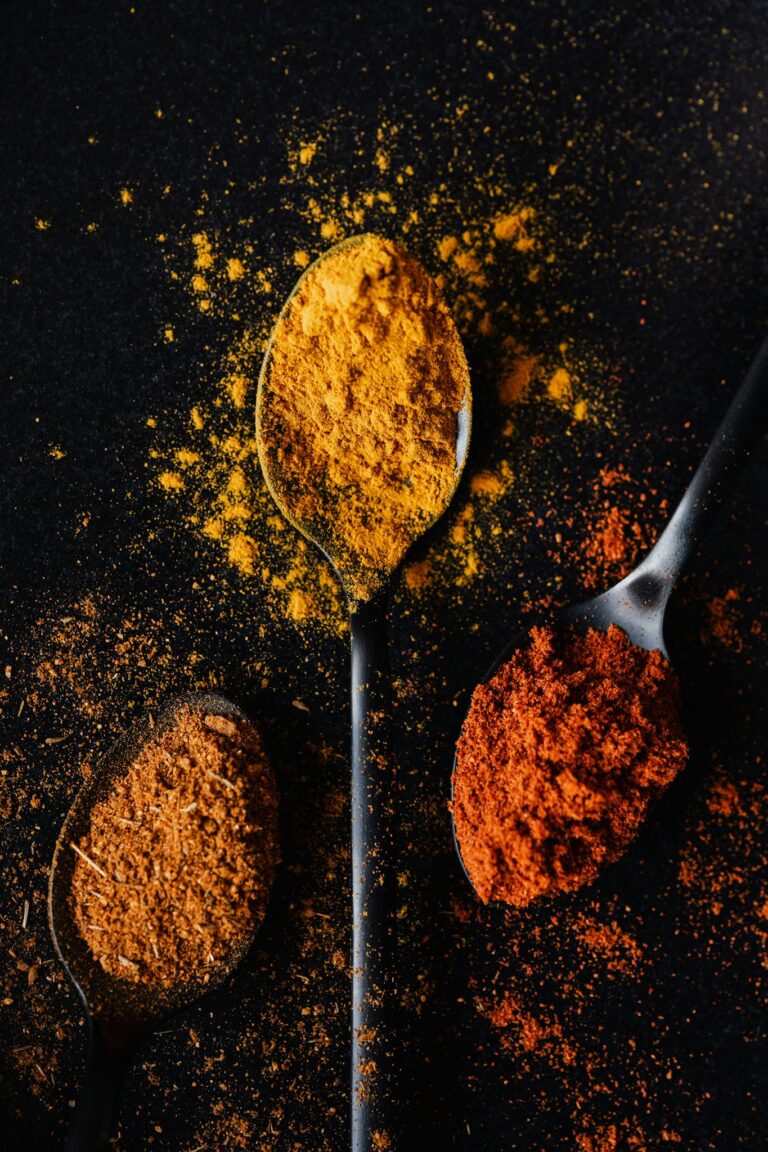 Overhead view of various colorful spices in spoons on a dark surface.